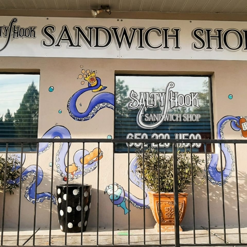 Exterior of Salty Hook Sandwich Shop with octopus mural, 'Now Open' sign, and potted plants.